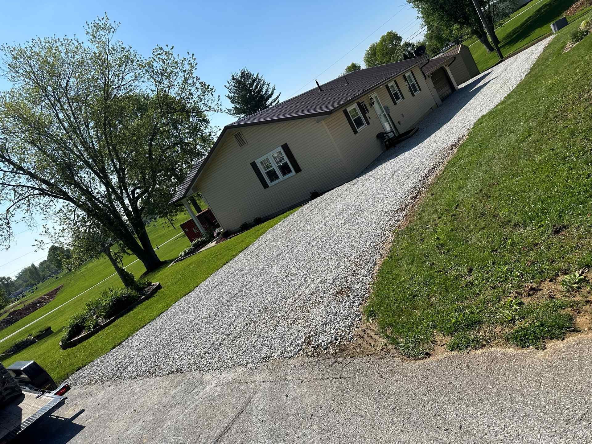 A tan single-story house with a dark roof and gravel driveway, surrounded by a green lawn and a large tree under a blue sky.