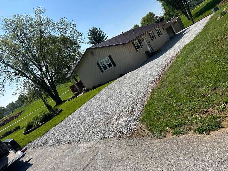 A beige single-story house with a dark roof sits behind a long gravel driveway, next to a large tree and grassy yard.
