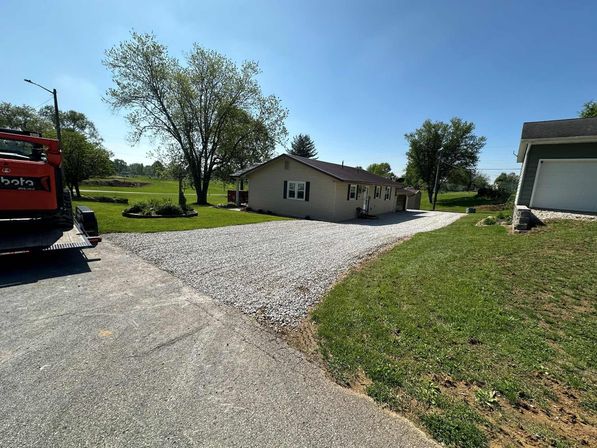 A beige house with a gravel driveway, seen on a sunny day with a corner of construction equipment in the foreground.