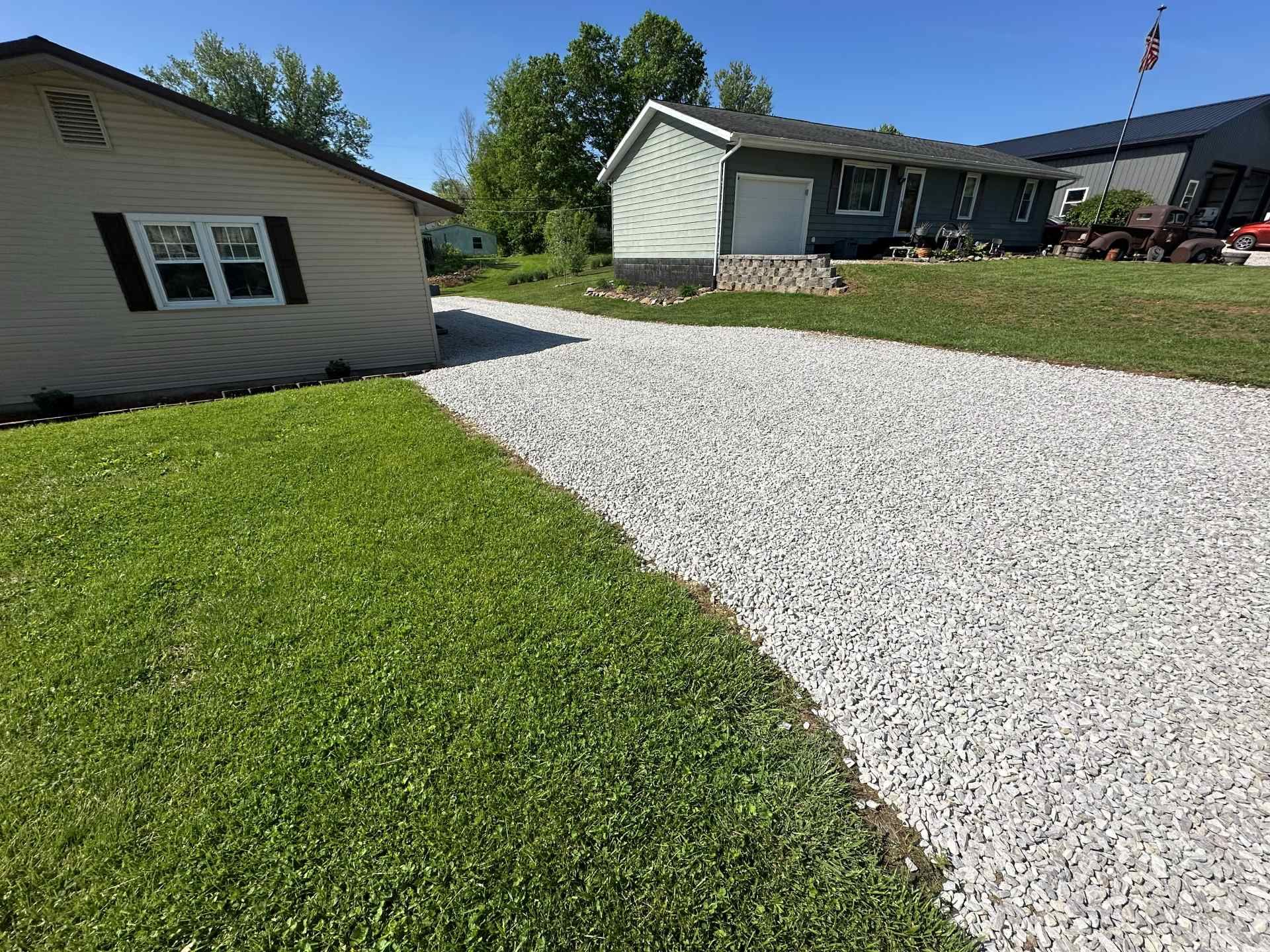 A gray gravel driveway leading toward a house on a sunny day with a green lawn in the foreground.