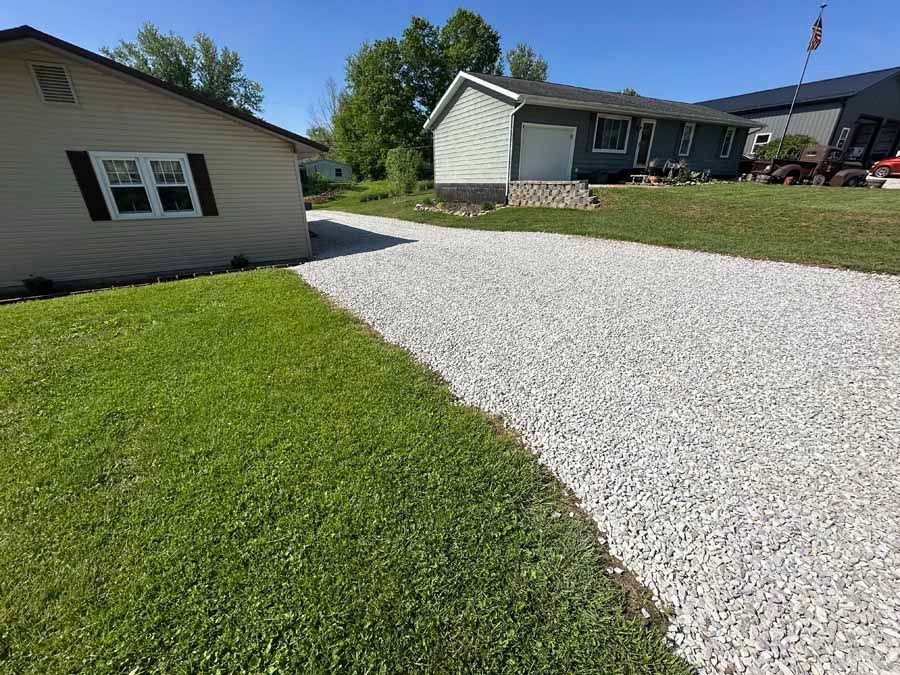 A wide view of a light-colored gravel driveway leading toward two houses on a sunny, clear day with a grassy lawn.