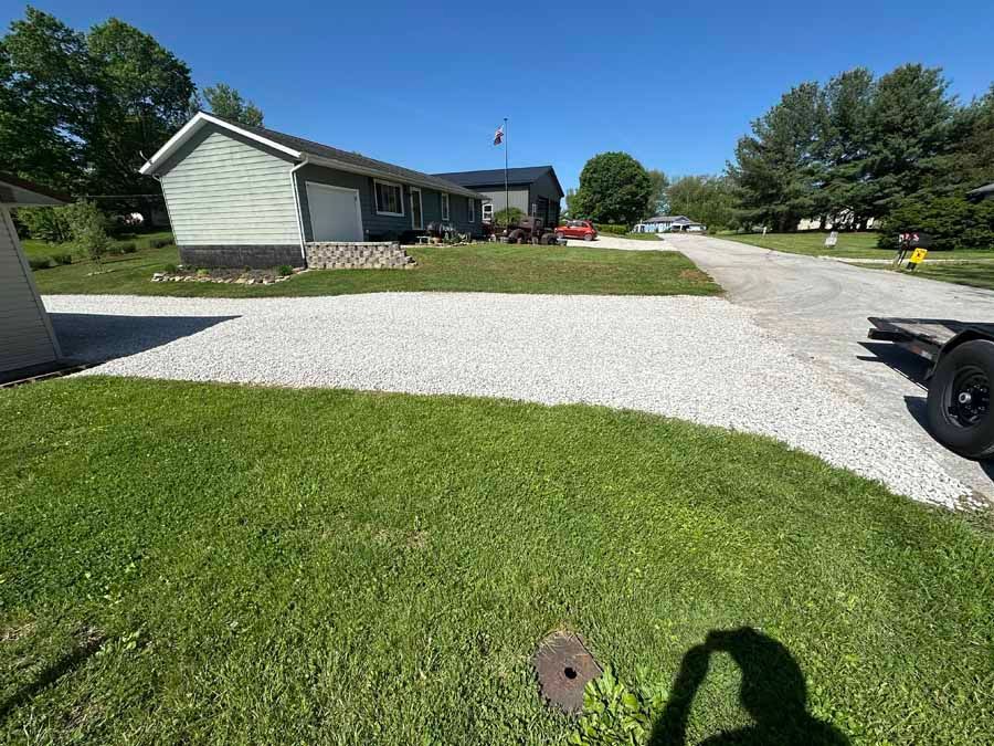 A light-colored house with a gravel driveway under a clear blue sky, with a portion of a trailer visible on the right.
