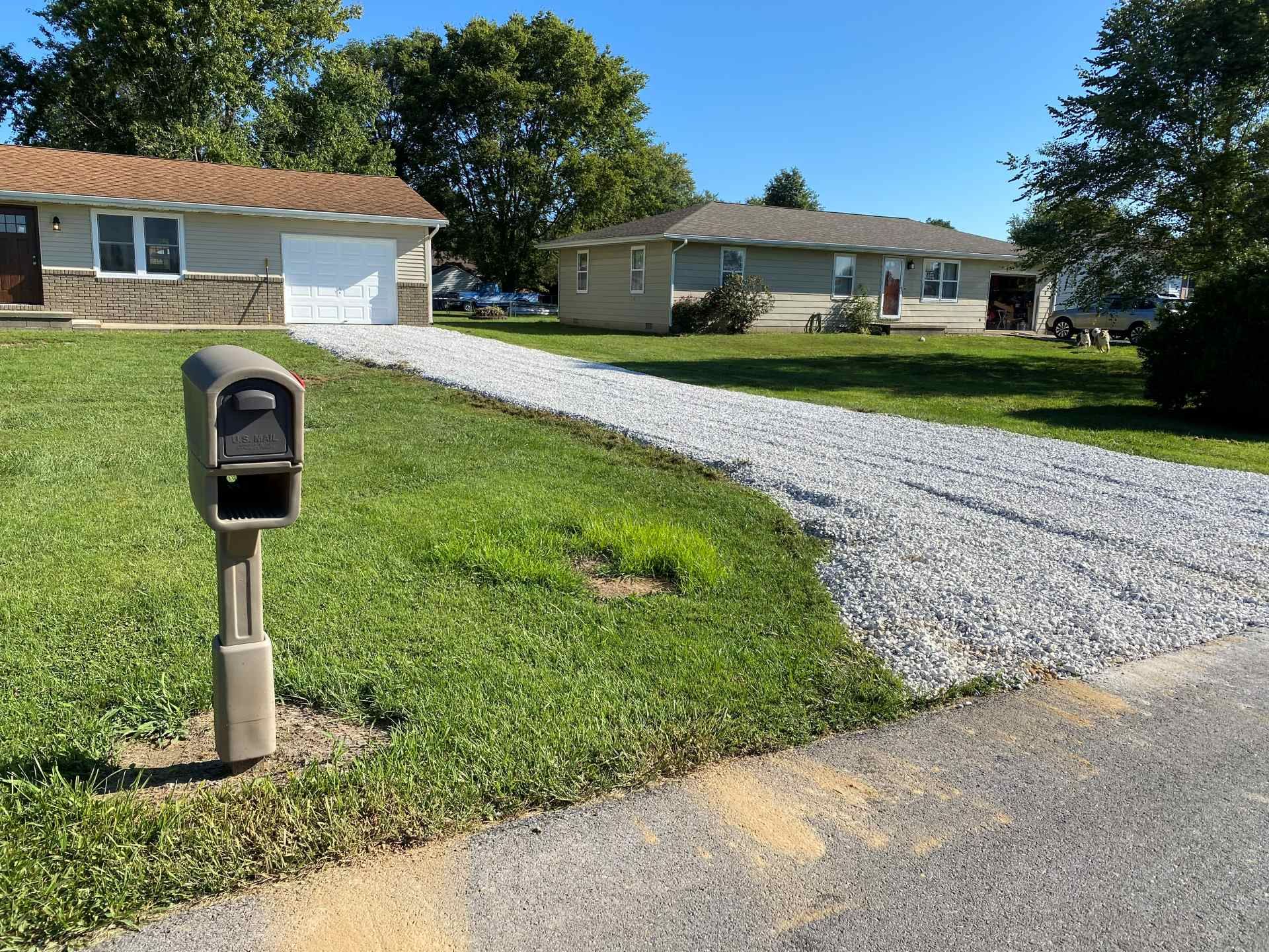 A rural suburban street scene showing a residential house, a gray gravel driveway, and a mailbox in the foreground.