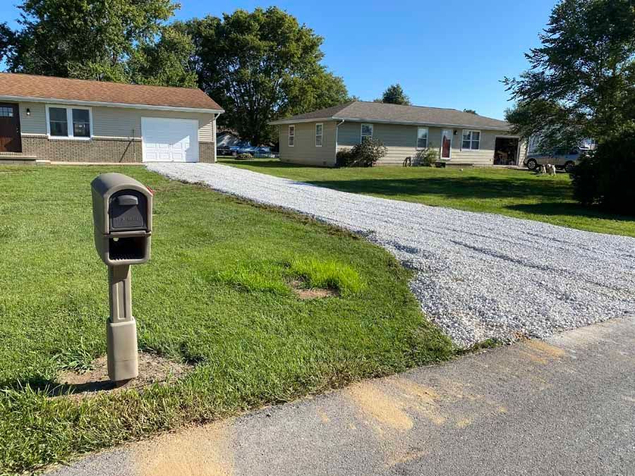 A beige house with a white garage door, featuring a gravel driveway and a pedestal mailbox in the front lawn.