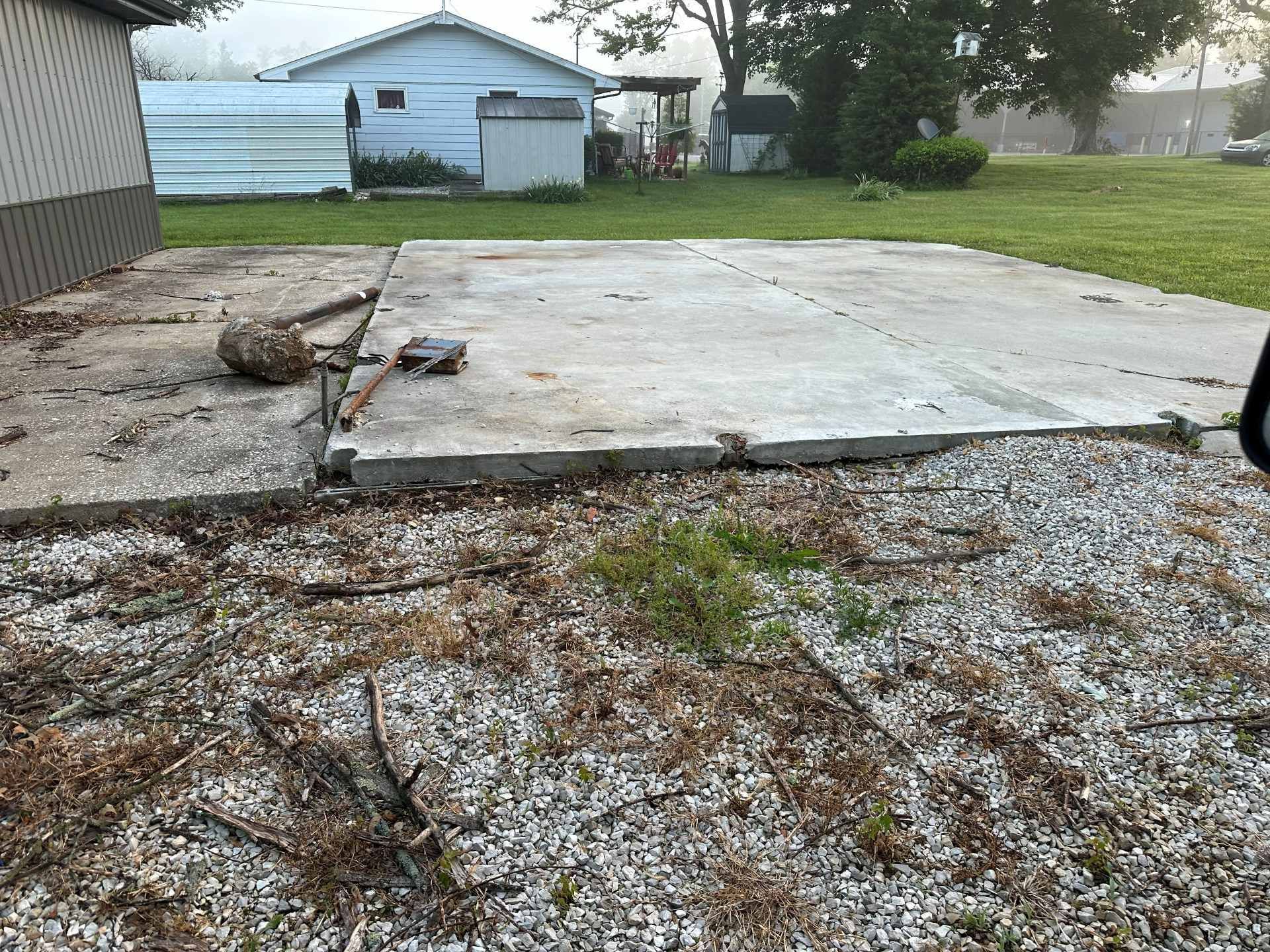 A concrete slab foundation sits in a yard near a house, with loose gravel and debris in the foreground.