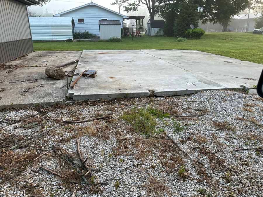 A concrete slab pad sits on a gravel and grass lot next to a building and storage sheds on a cloudy day.
