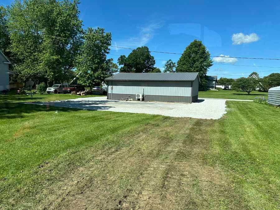 A detached grey garage sits on a gravel driveway in a grassy yard under a clear blue sky.