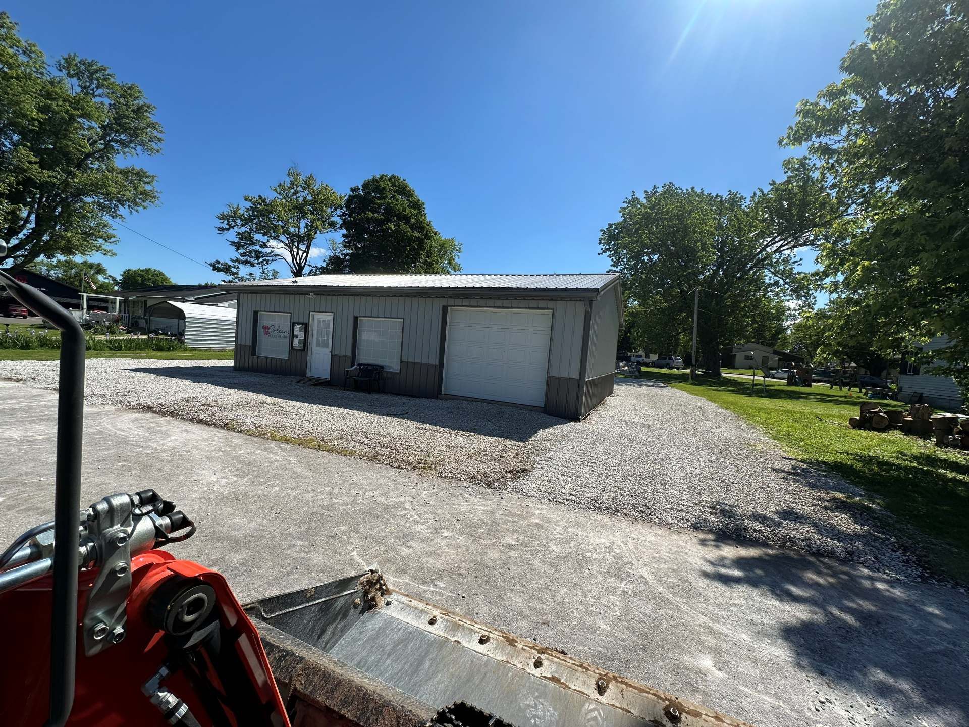 A grey metal shed with white garage doors stands on a gravel lot under a clear blue sky, viewed from a loader bucket.