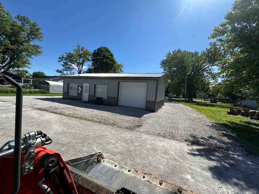 View from a front loader bucket looking at a light gray, single-story metal building on a sunny day.