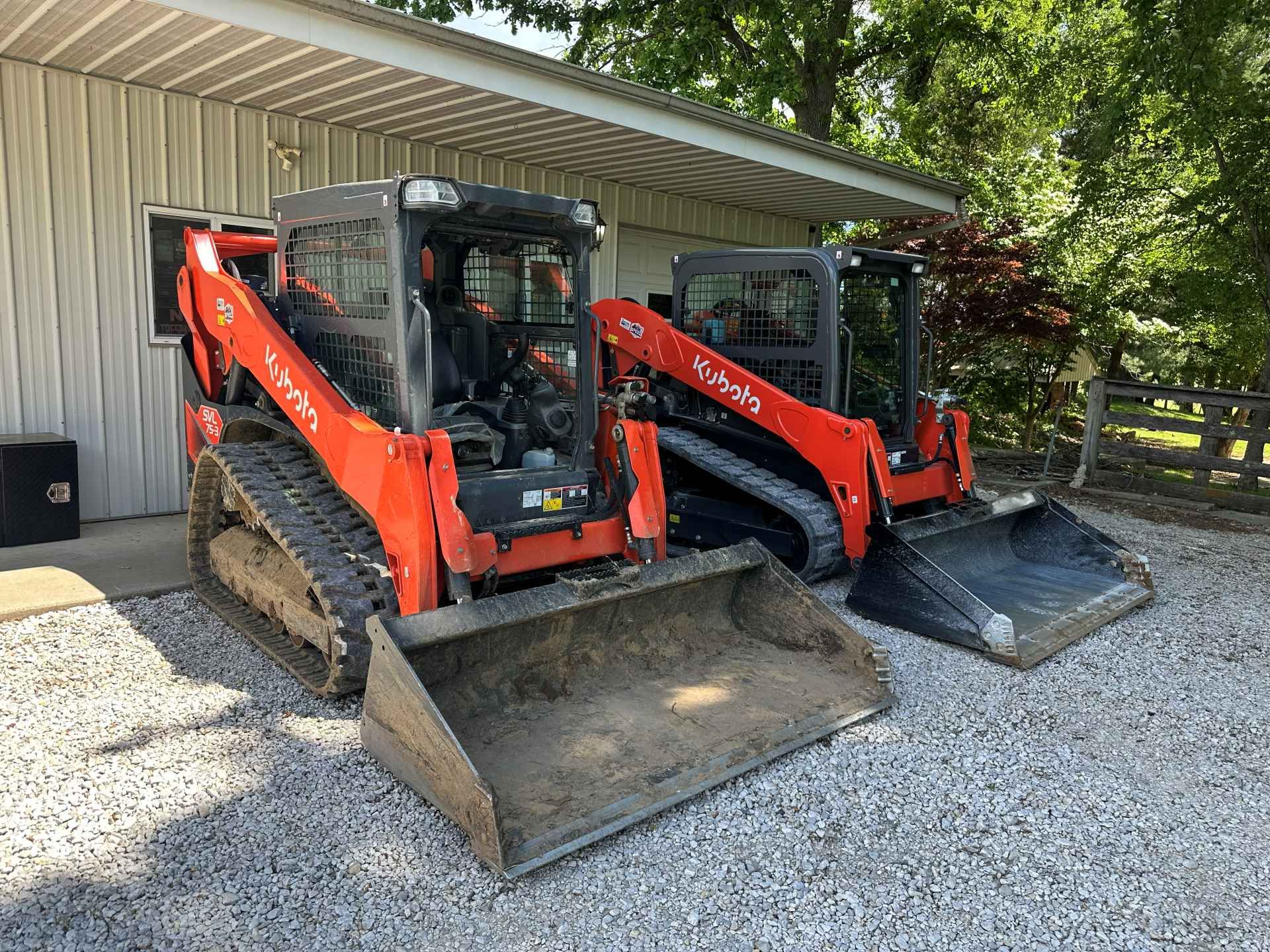 Two bright orange Kubota skid-steer loaders with tracks parked on a gravel driveway in front of a metal building.