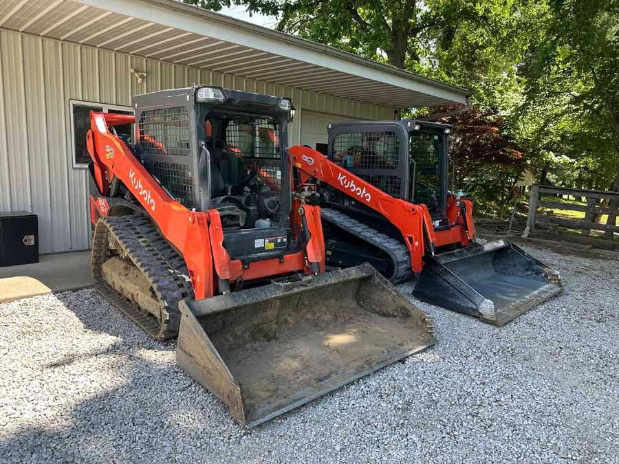 Two orange Kubota compact track loaders parked side-by-side on a gravel driveway in front of a metal building.