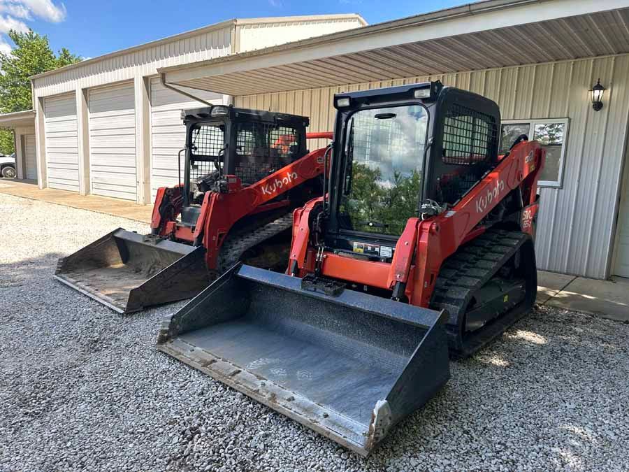 Two orange Kubota compact track loaders parked side-by-side on a gravel lot in front of a metal building.