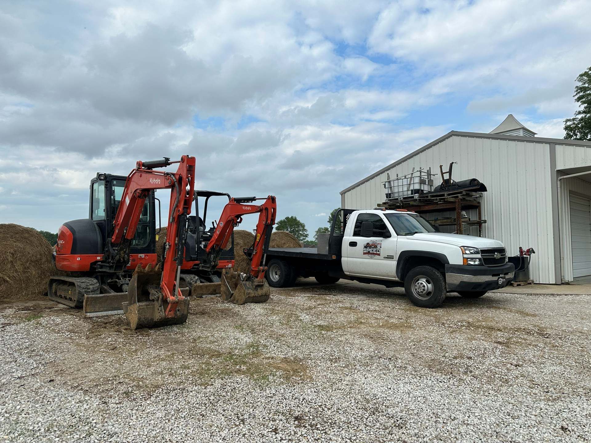 Two orange excavators parked on gravel next to a white pickup truck in front of a metal workshop under a cloudy sky.