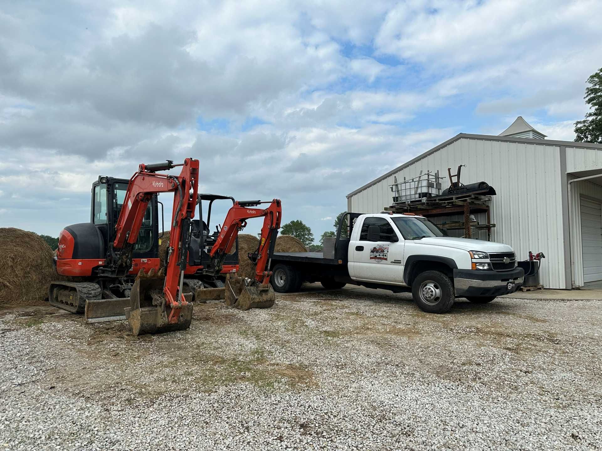 Two orange construction excavators parked next to a white flatbed truck in front of a metal warehouse.