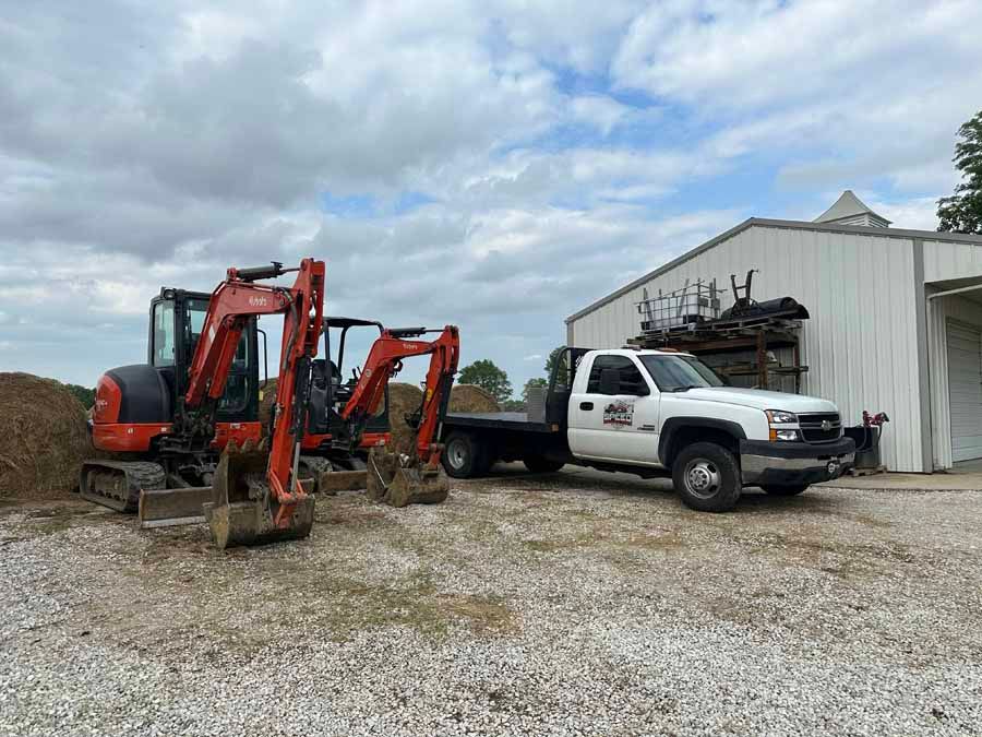 Two orange excavators parked next to a white flatbed truck in front of a metal building on a gravel lot.