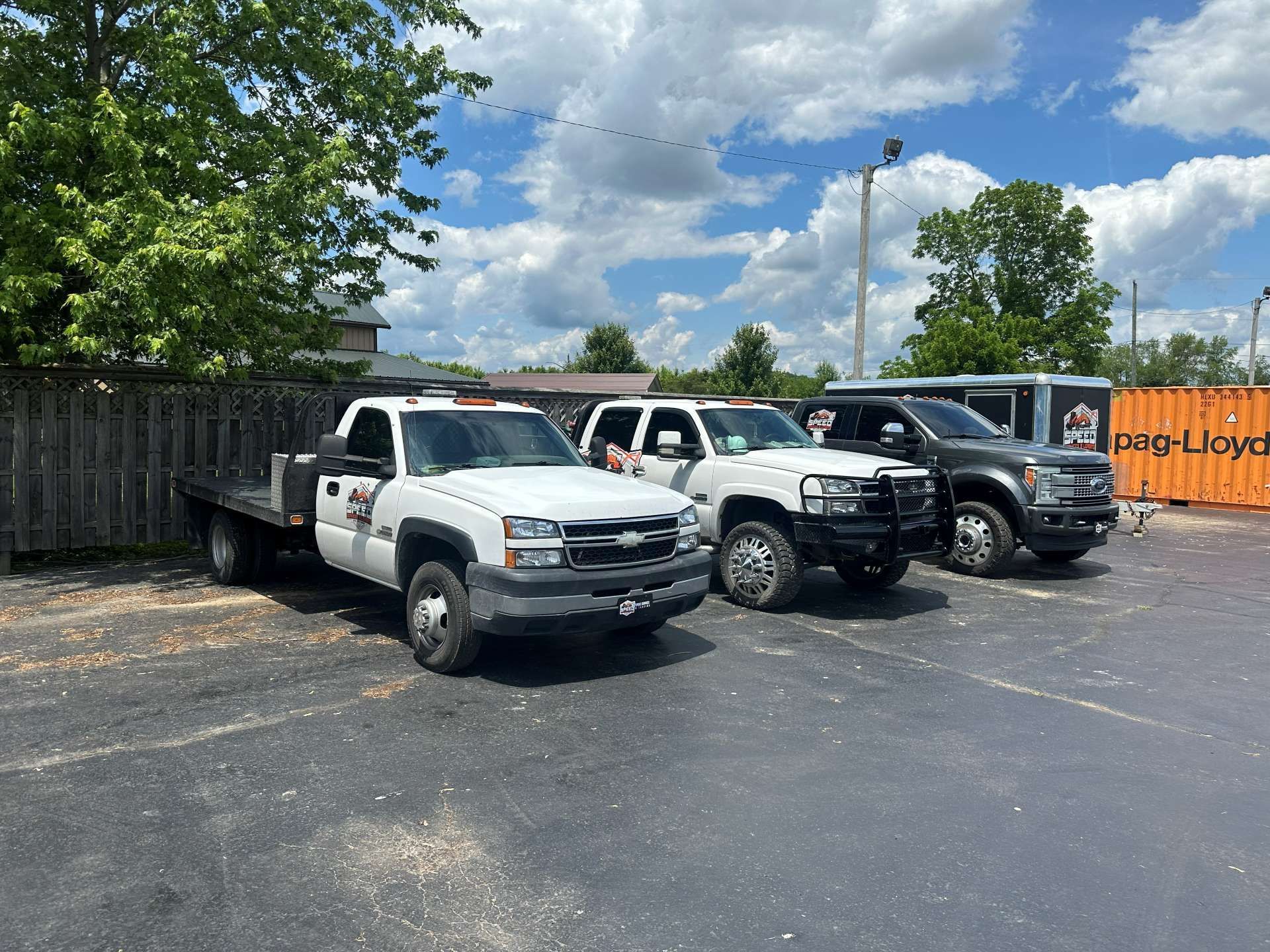 Three white and dark pickup trucks parked in a lot next to a wooden fence and an orange shipping container.