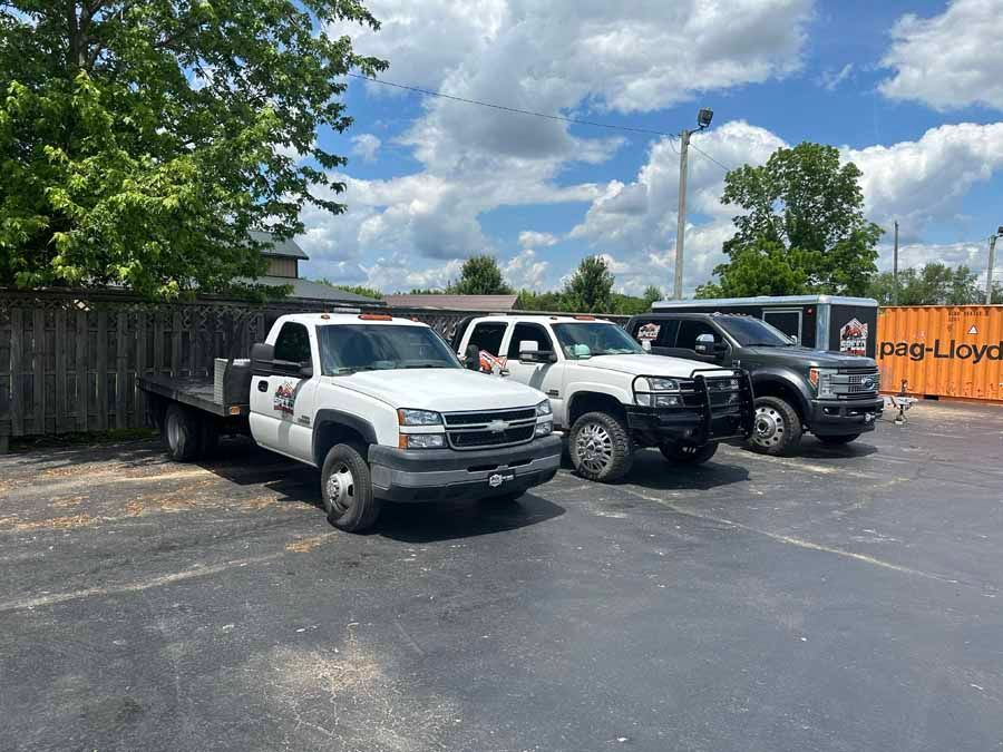 Three trucks, two white and one dark grey, are parked in a row on an asphalt lot near a wooden fence and shipping container.
