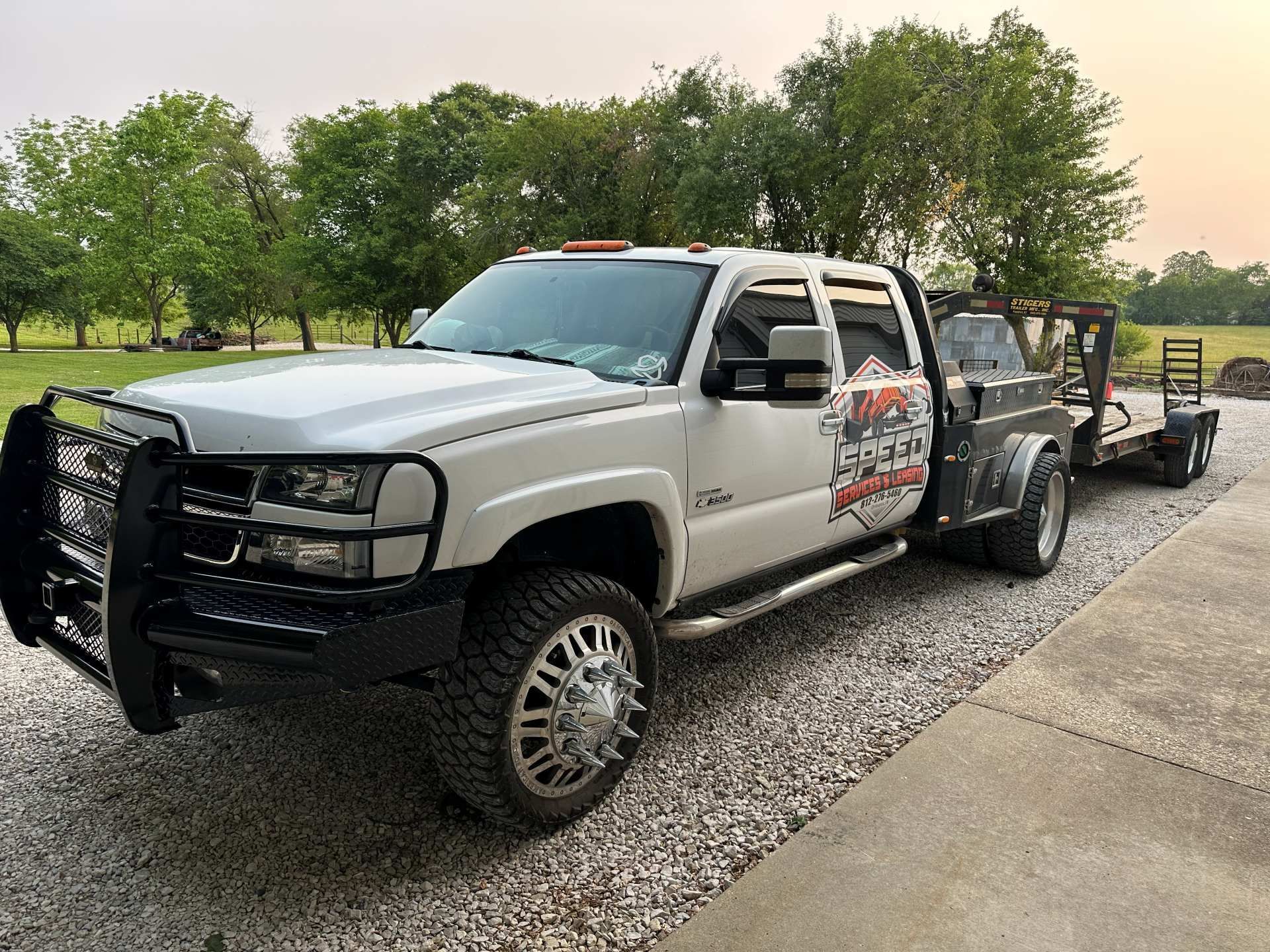 A white heavy-duty pickup truck with a black brush guard and flatbed, parked on gravel, pulling a utility trailer.
