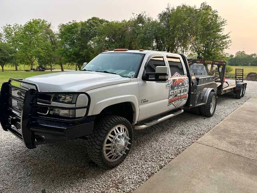 White heavy-duty crew cab dually pickup truck with a black brush guard, parked on a gravel lot hitched to a flatbed trailer.