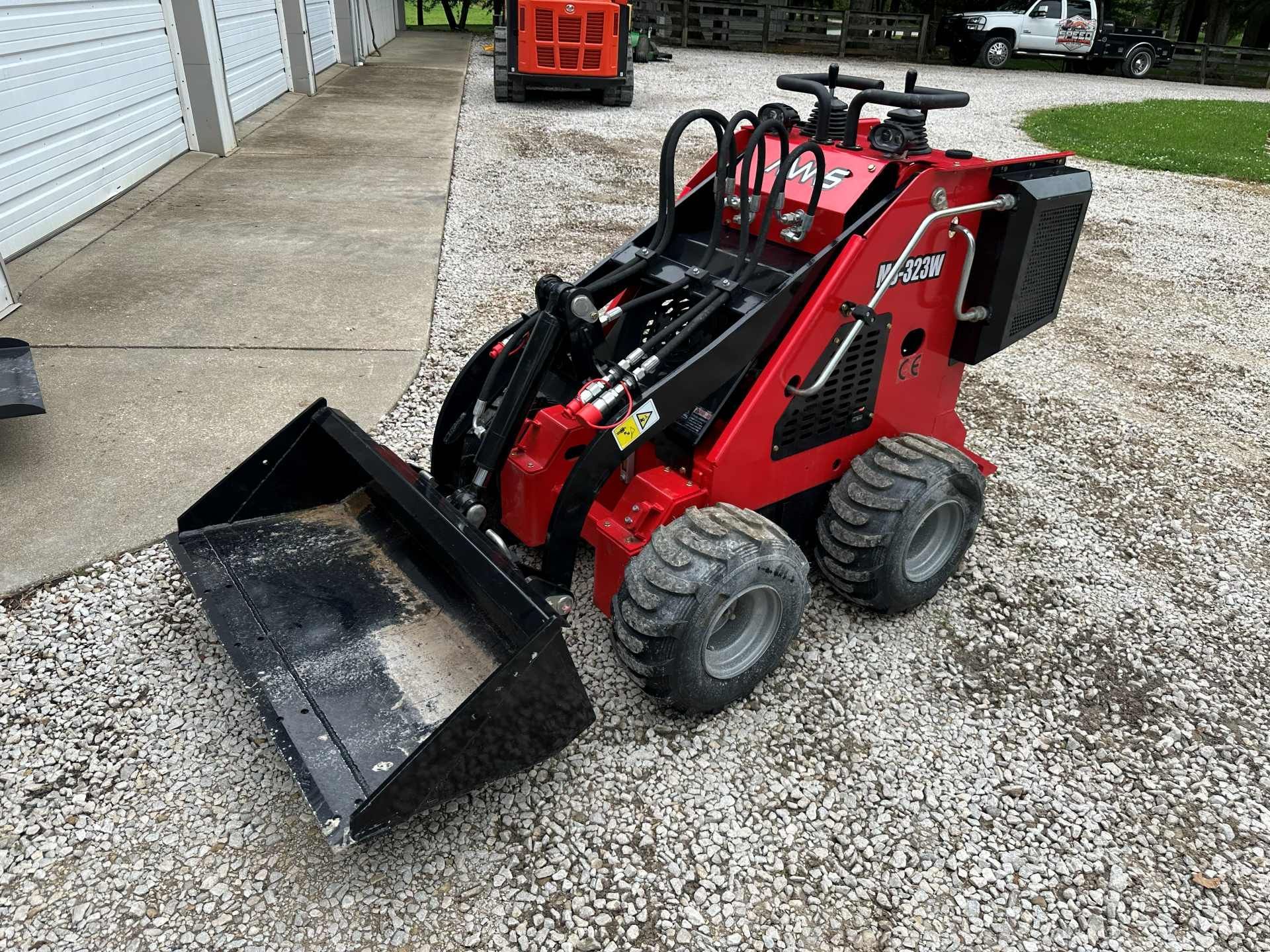 A red stand-on compact utility loader with a black front bucket parked on a gravel surface.