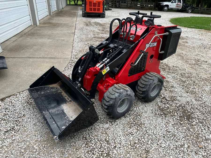 A compact red stand-on skid steer loader with a front bucket parked on a gravel surface.