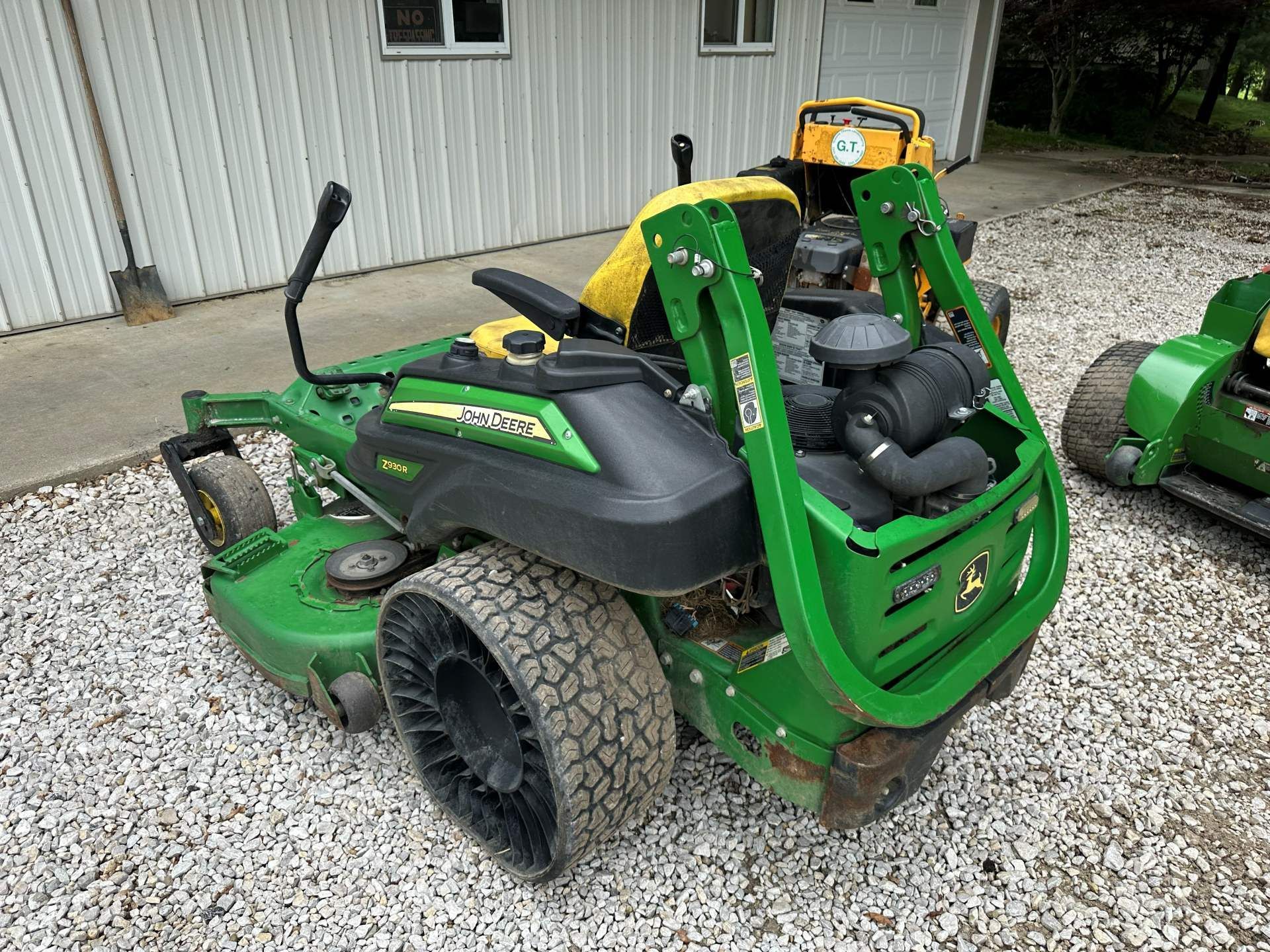 A green and yellow John Deere ZTrak zero-turn mower parked on a gravel surface next to a white building.