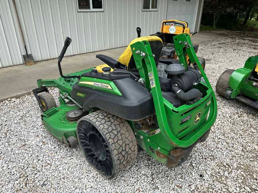A green and yellow John Deere zero-turn lawn mower parked on a gravel surface next to a white metal building.