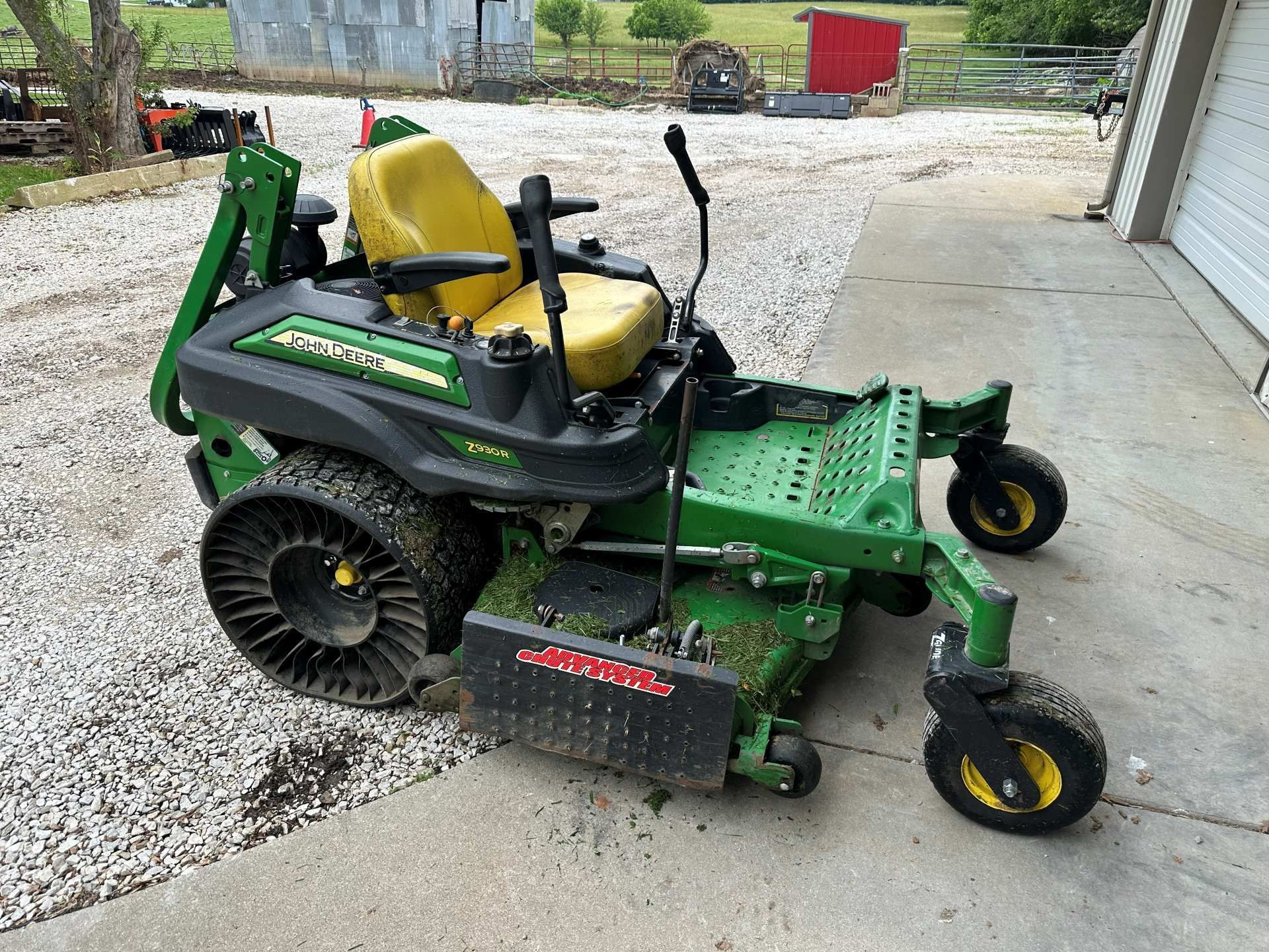 A green and black John Deere zero-turn lawn mower with yellow seating parked on a gravel driveway near a rural building.
