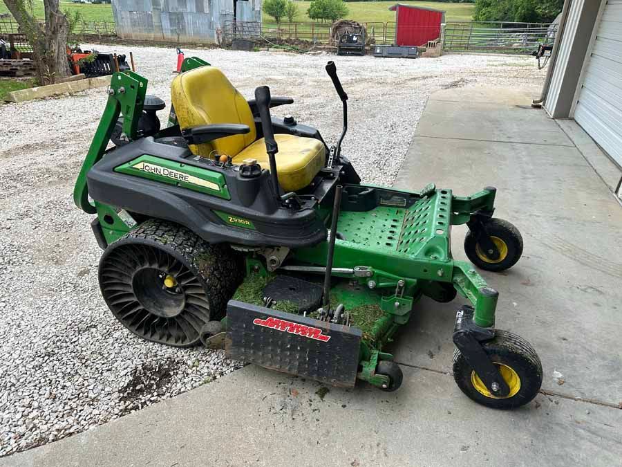 A green and yellow John Deere ZTrak zero-turn mower parked on a gravel driveway next to a building.