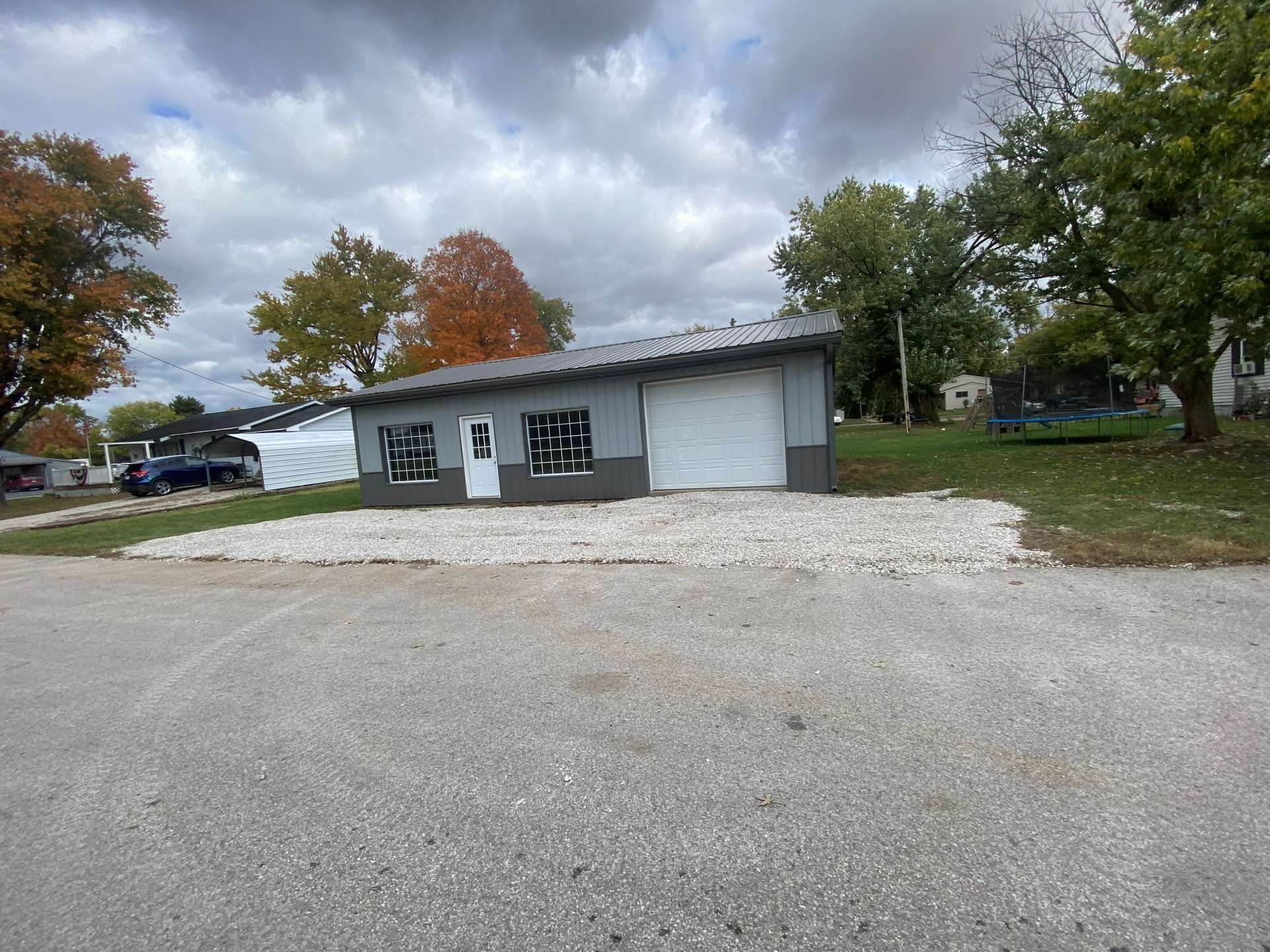A gray, single-story garage with a white door and two windows sits on a gravel lot under a cloudy sky.