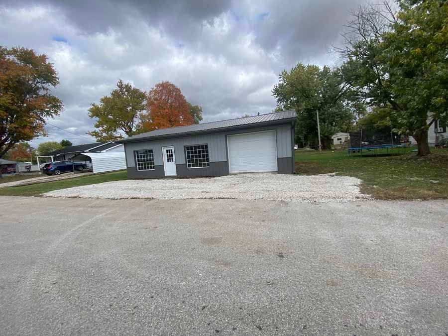 A gray, one-story metal building with a white garage door, a side door, and two windows, set on a gravel lot near trees.