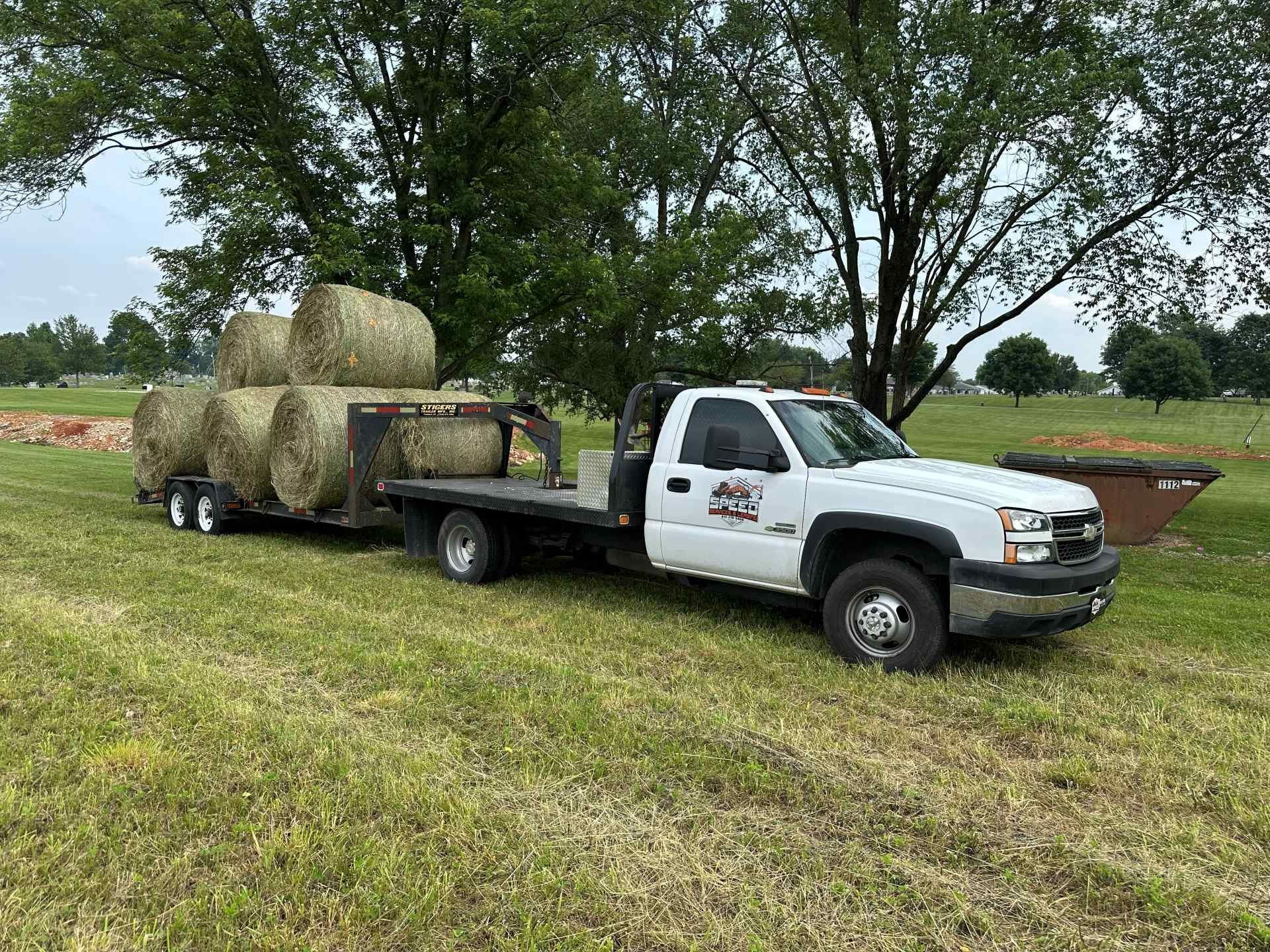 A white flatbed truck hauling a trailer loaded with six round hay bales in a grassy field under a cloudy sky.