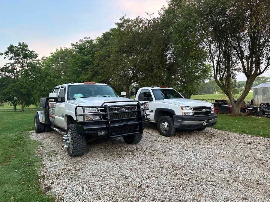 Two white Chevrolet dually pickup trucks parked side-by-side on a gravel driveway near trees.