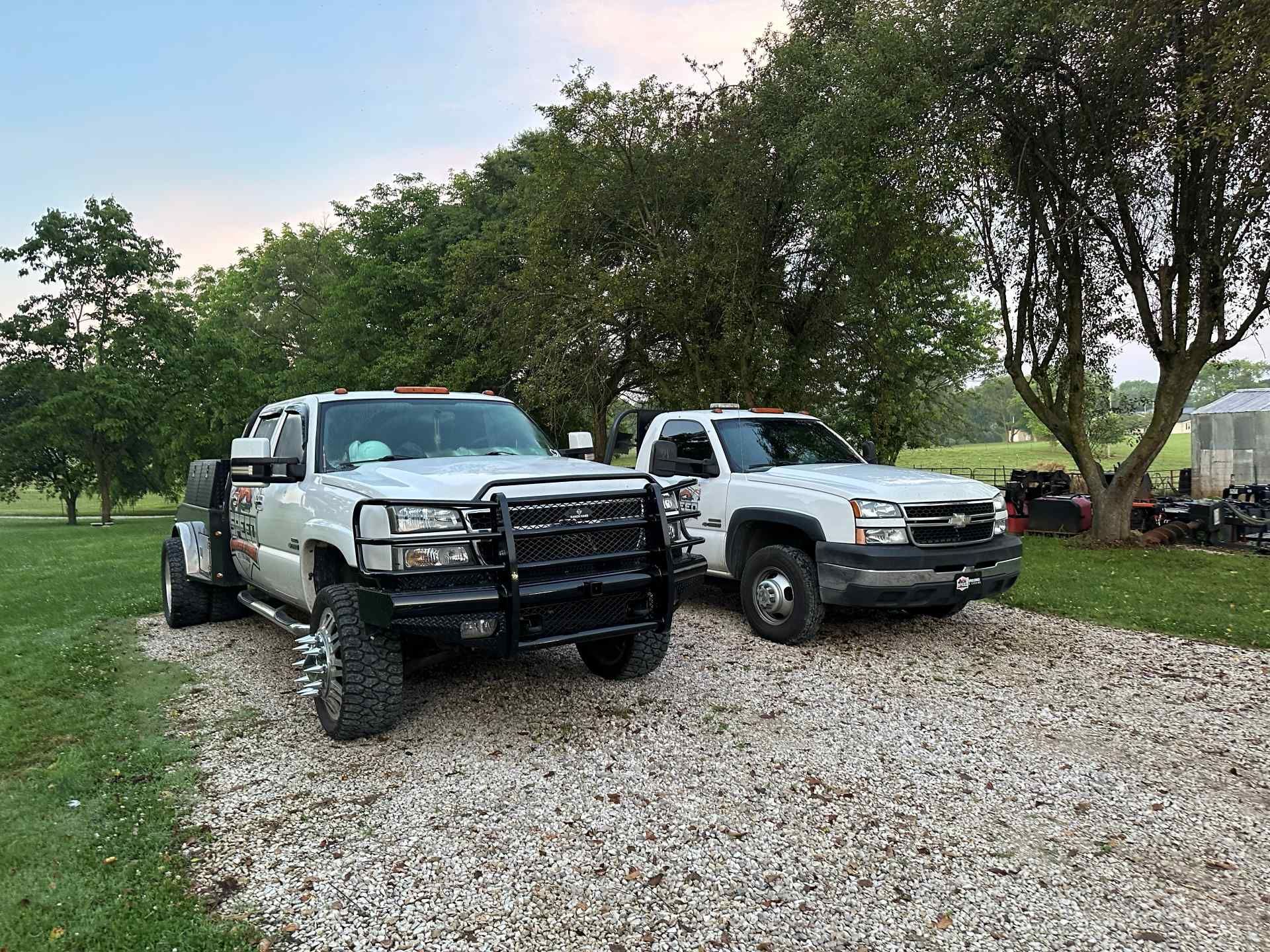 Two white Chevrolet pickup trucks parked on a gravel lot near trees during the daytime.
