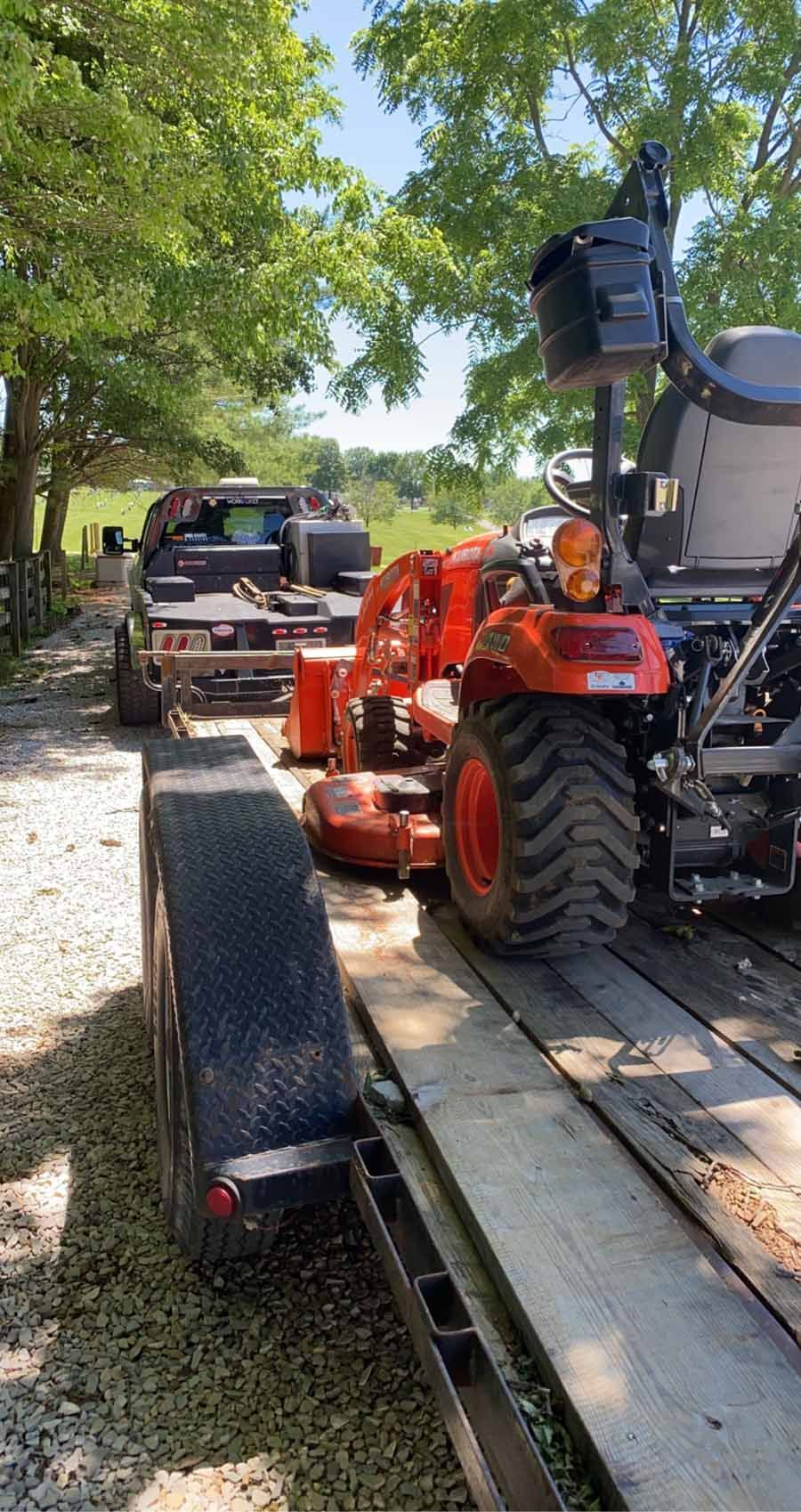 An orange tractor loaded onto a flatbed trailer on a gravel road, pulled by a truck in a sunny outdoor setting.
