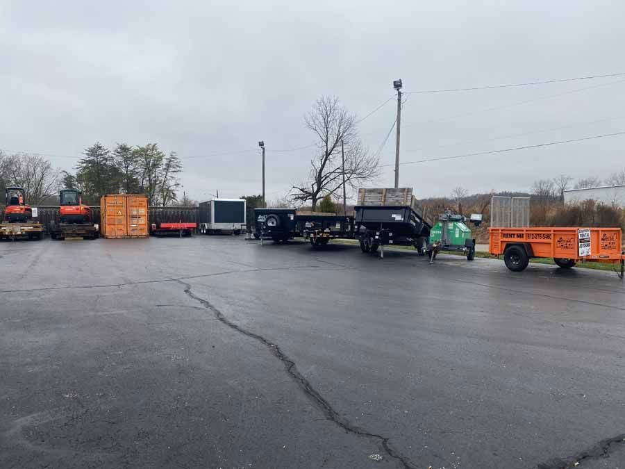 A row of construction equipment, including excavators, storage containers, and trailers, parked on an asphalt lot.