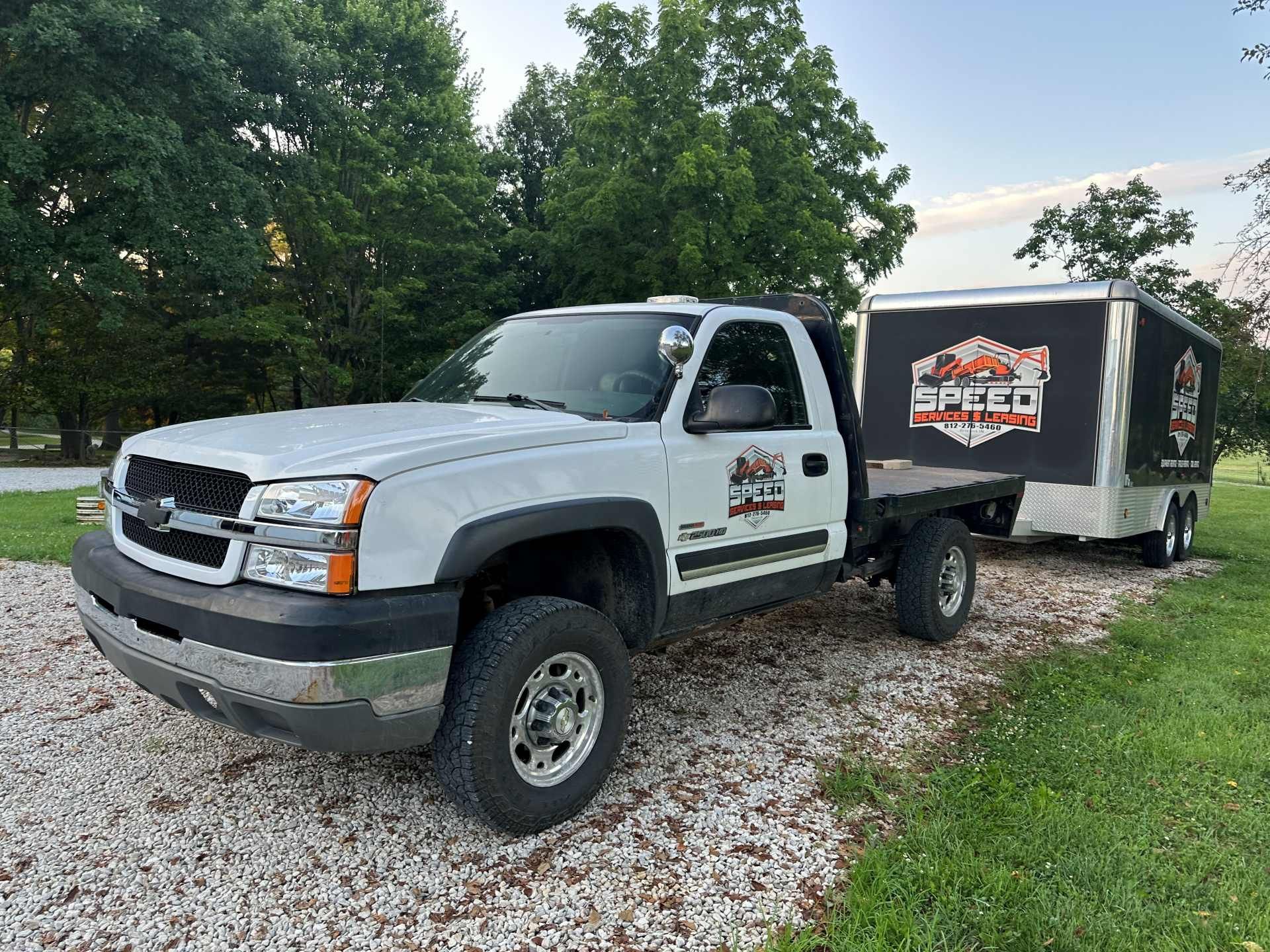 A white Chevrolet flatbed truck with a matching branded trailer parked on a gravel lot near trees.