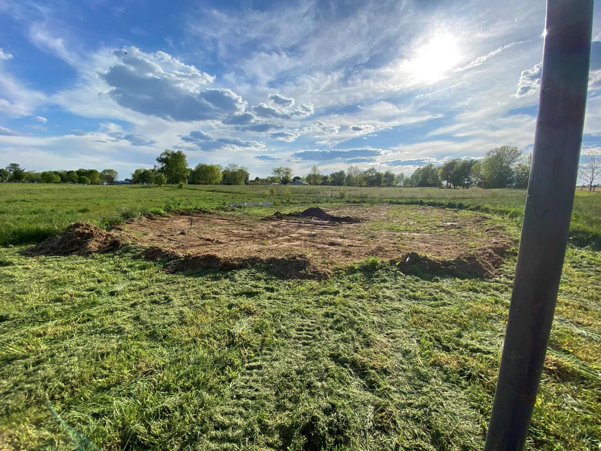 A circular patch of tilled, brown earth sits in a bright green field under a sunny, blue sky with scattered clouds.