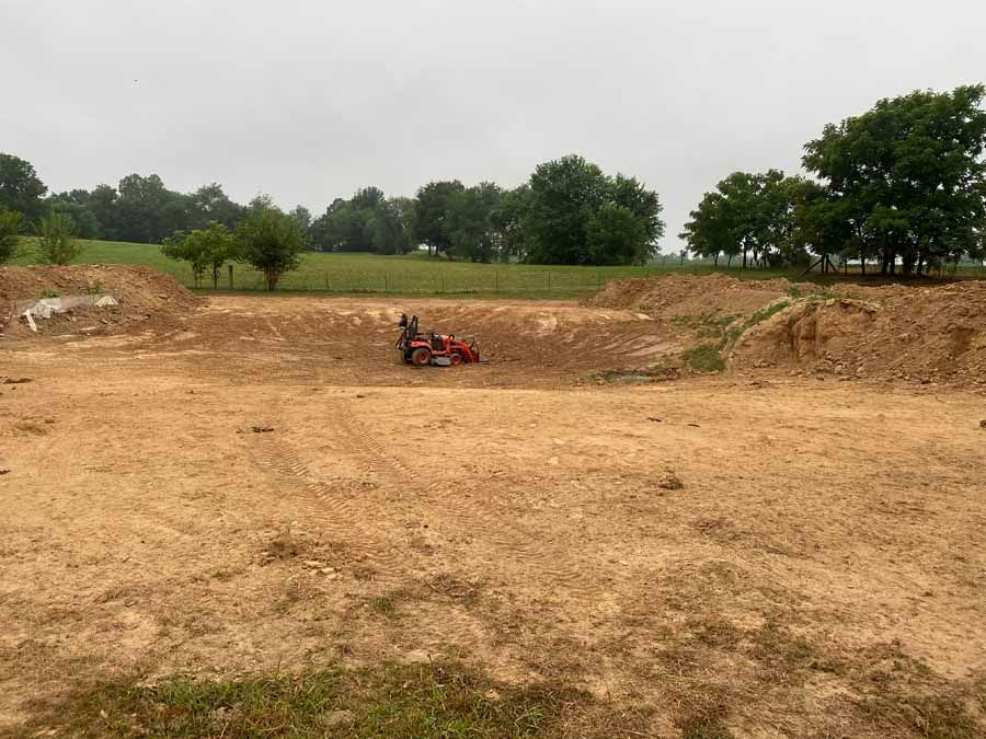 A red mower sits in the center of a large, excavated dirt lot against a backdrop of trees under an overcast sky.