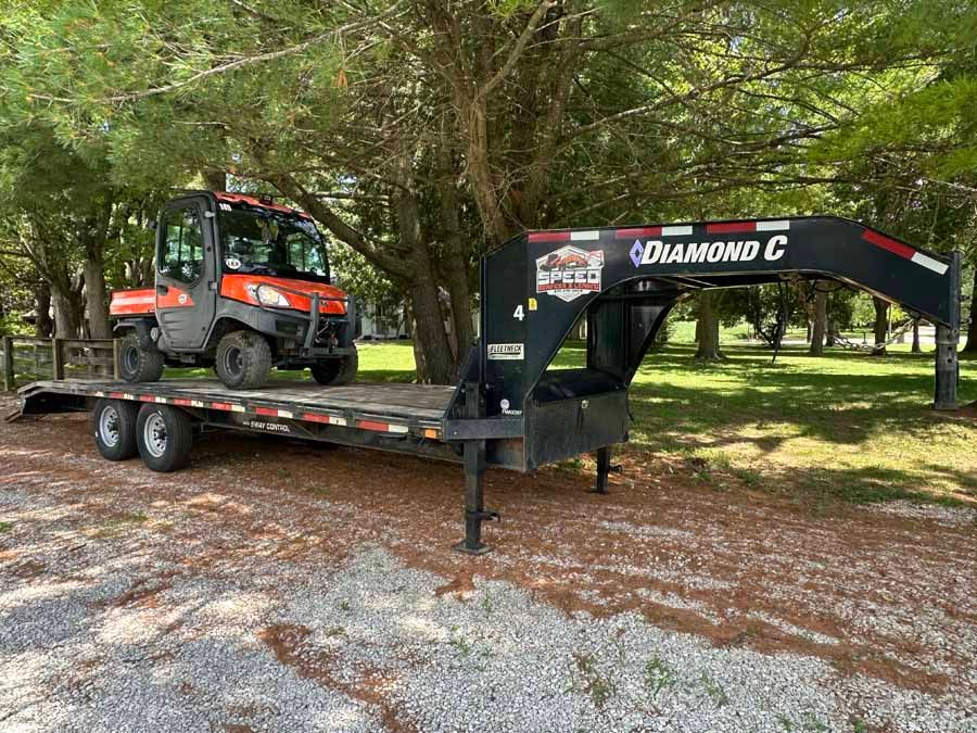 An orange utility vehicle parked on a black Diamond C flatbed gooseneck trailer outdoors on a gravel drive.