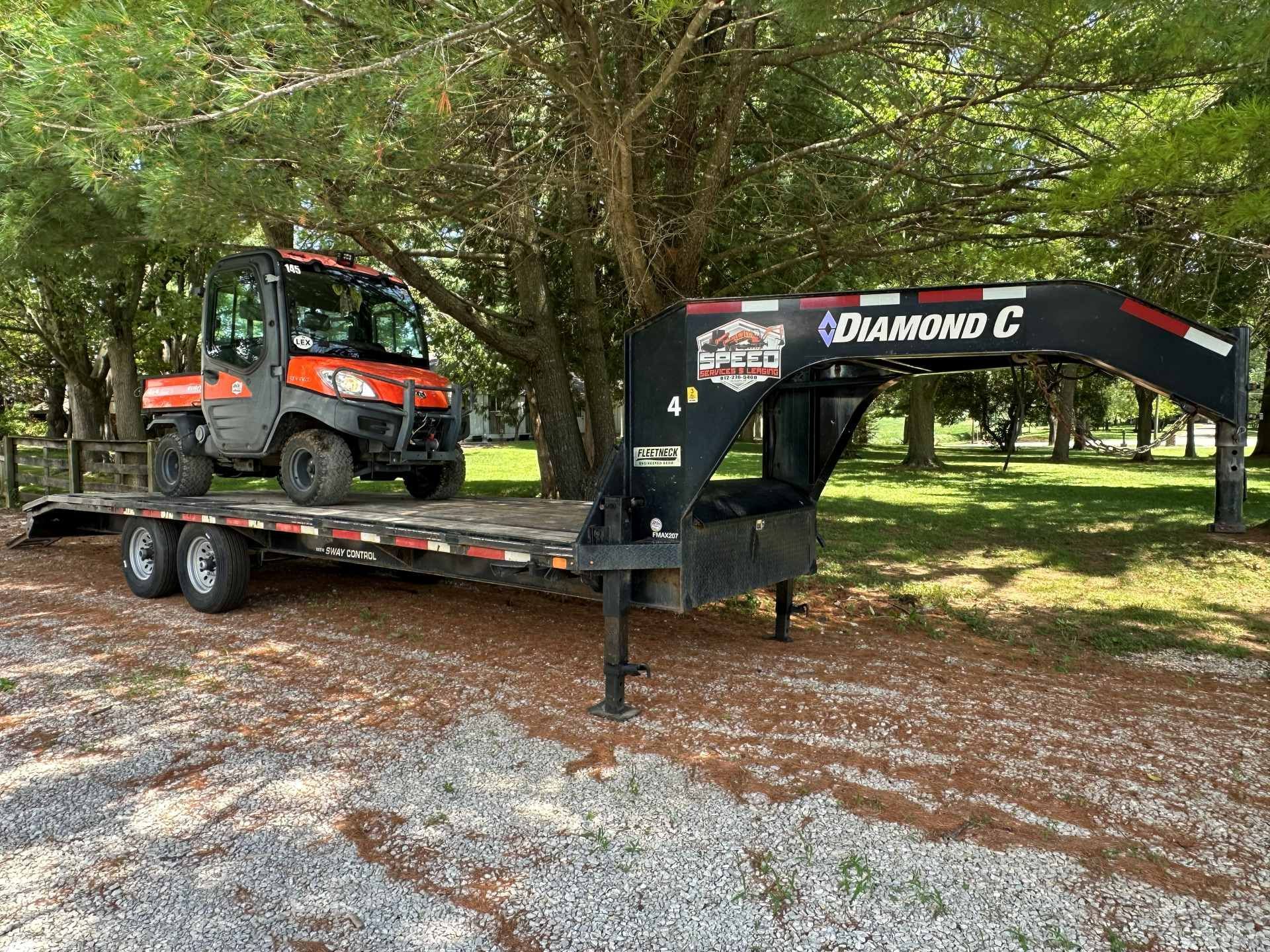 An orange skid steer loader sits on a black Diamond C gooseneck trailer parked on a gravel surface under leafy trees.