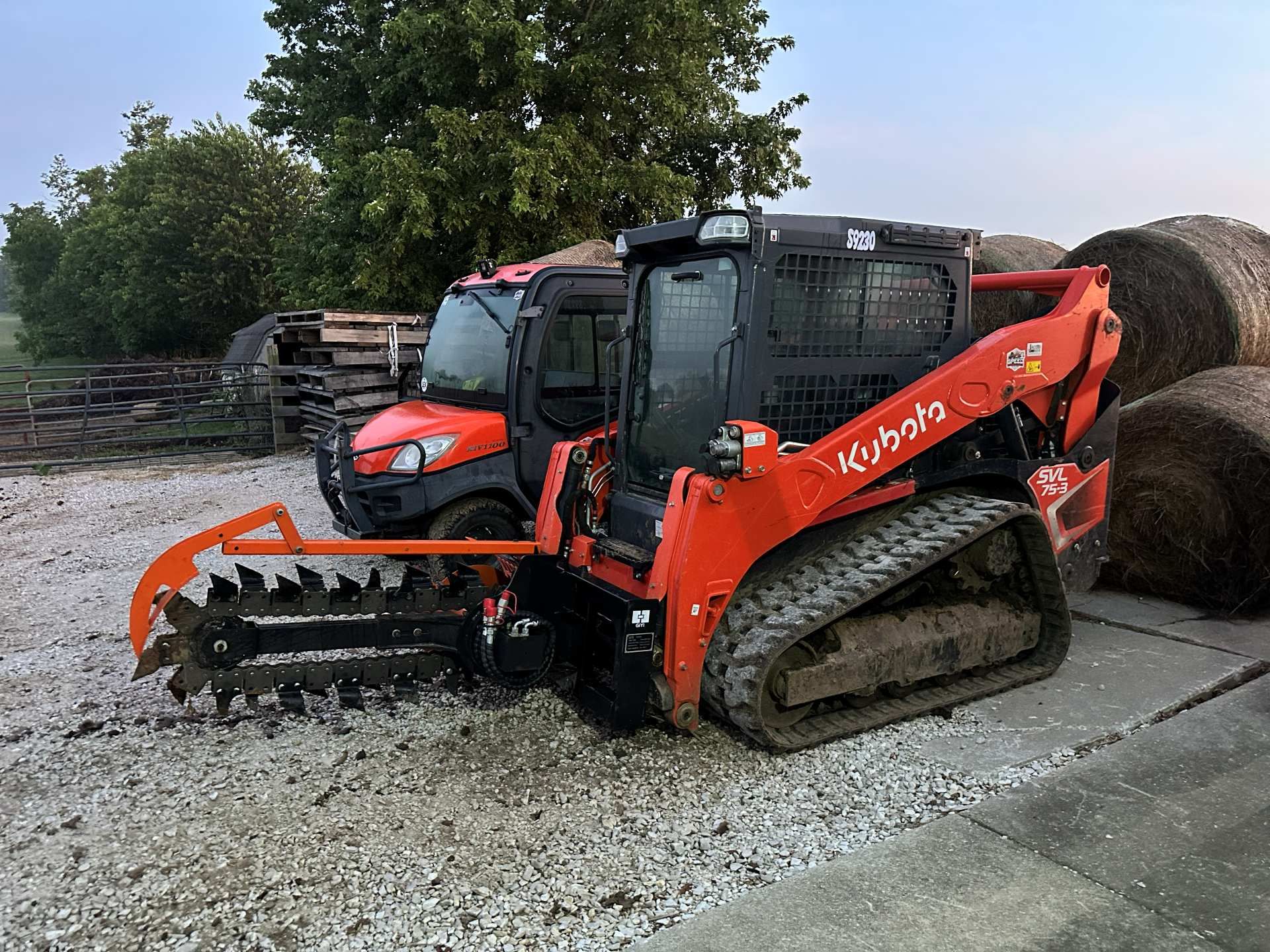 A bright orange Kubota tracked skid steer equipped with a trencher attachment, parked outdoors next to hay bales.