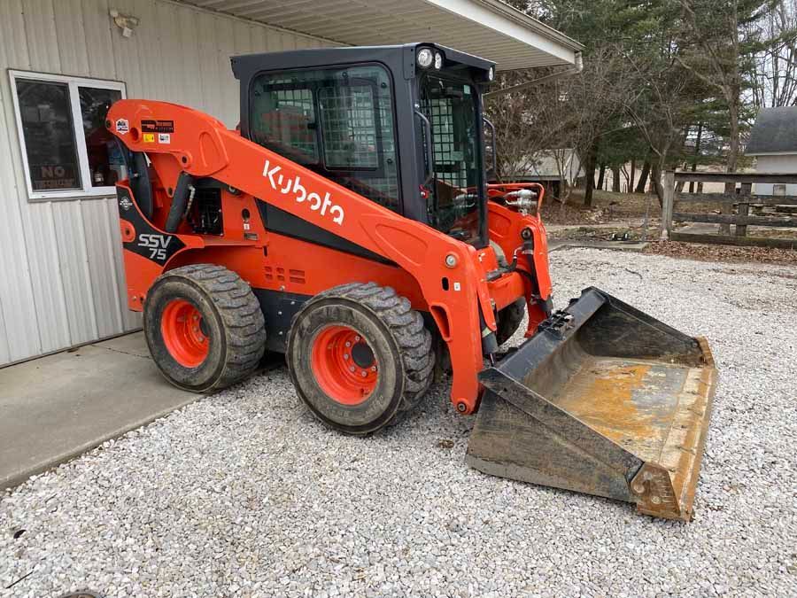 An orange Kubota SSV75 skid steer loader with a front bucket parked on a gravel lot next to a white building.