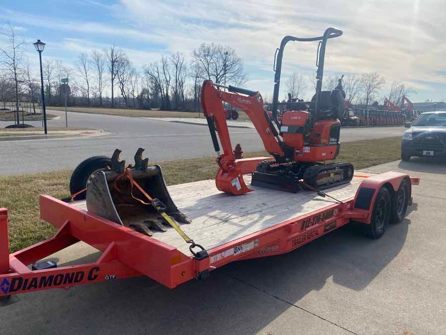 An orange Kubota excavator and a detached shovel bucket are secured to a red Diamond C flatbed trailer outdoors.