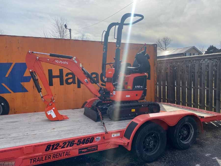 An orange Kubota mini excavator secured on a red flatbed utility trailer parked outdoors.
