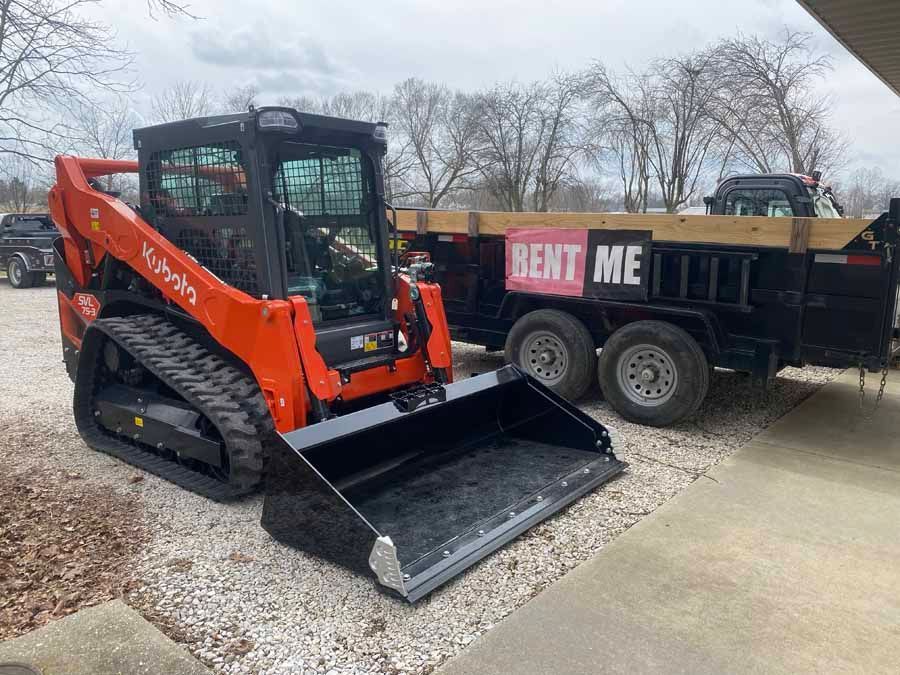 A bright orange Kubota track loader with a large black bucket parked next to a black dump trailer labeled 