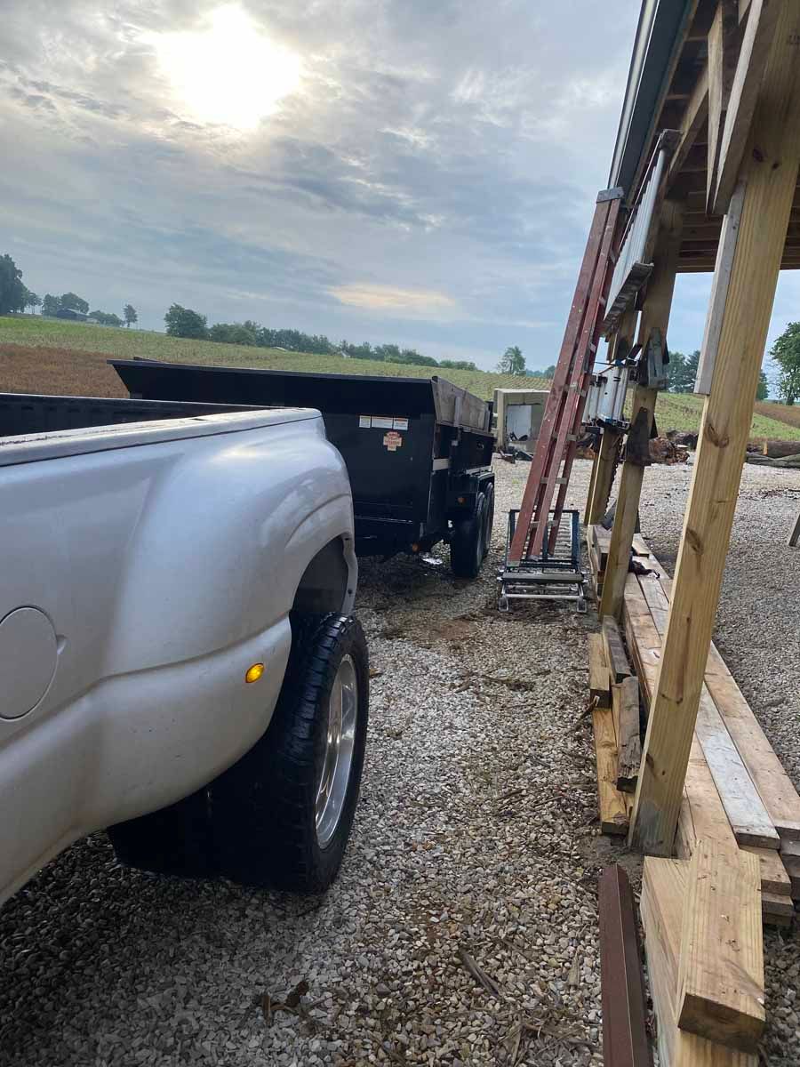 A white dually truck parked next to a black dump trailer on a gravel lot under a wooden structure with a leaning ladder.