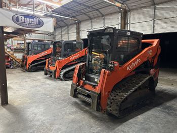 Three orange Kubota tracked skid steer loaders parked in a line inside a warehouse.