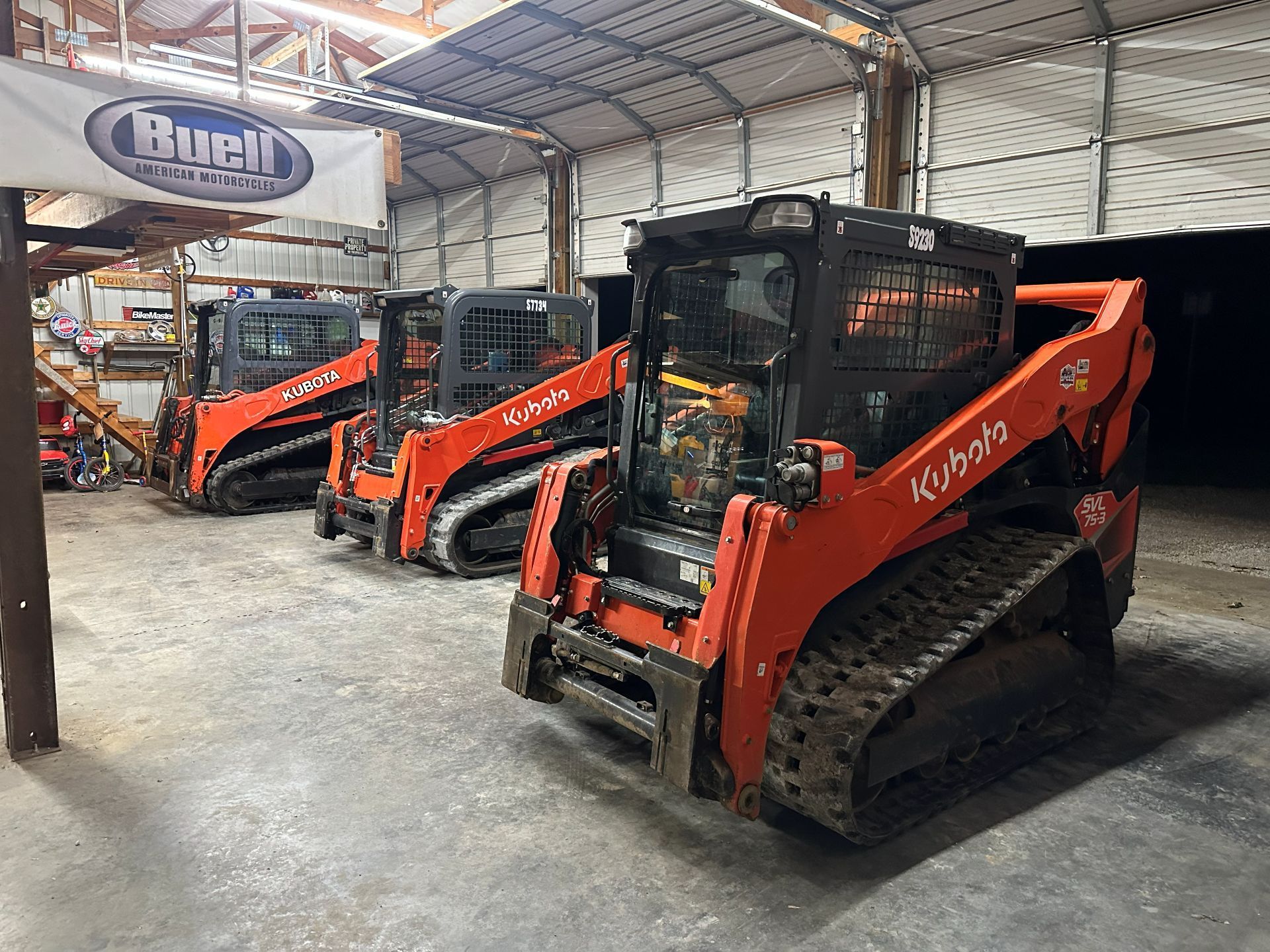 Three orange Kubota tracked skid steer loaders parked in a line inside an industrial warehouse.
