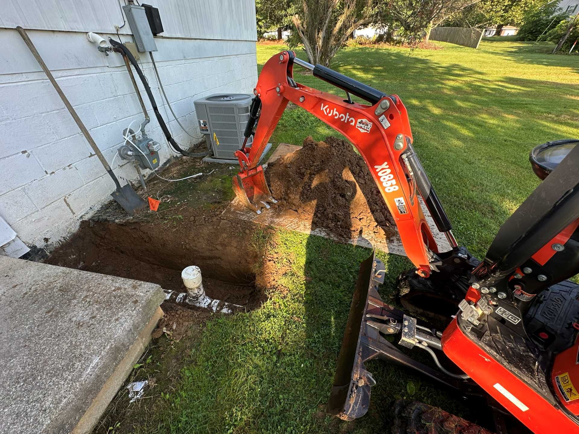 An orange mini excavator digs a trench in a residential yard next to a white house foundation and an HVAC unit.